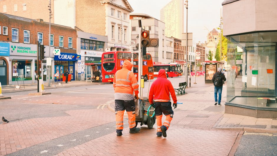 Two city street cleaners wearing high-visibility orange and red jackets with reflective strips are performing surface cleaning and maintenance in a busy area of Brentford. They are pushing a trolley w
